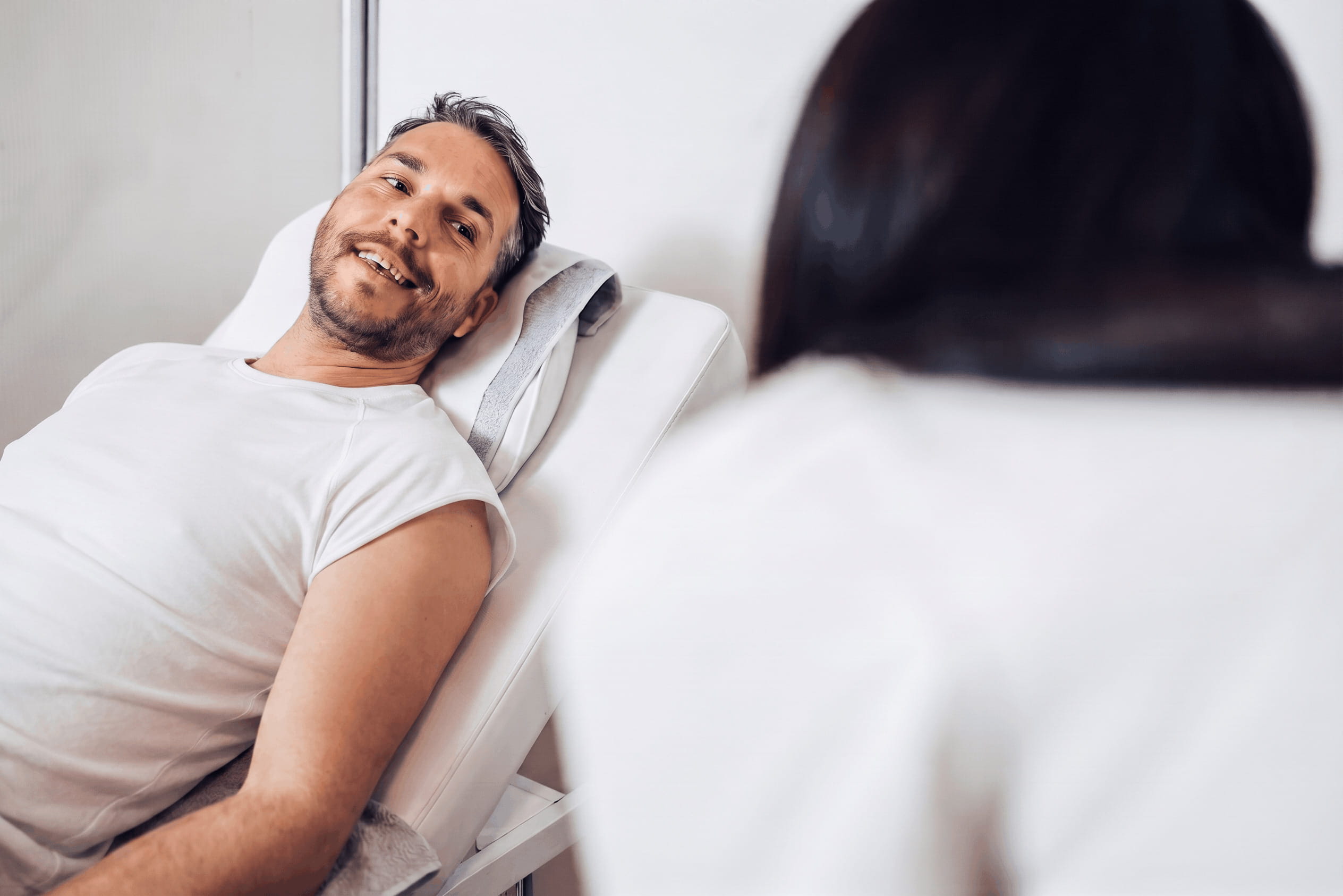 A smiling man in a white t-shirt lies on a medical examination table, looking at a person with dark hair whose back is to the camera.