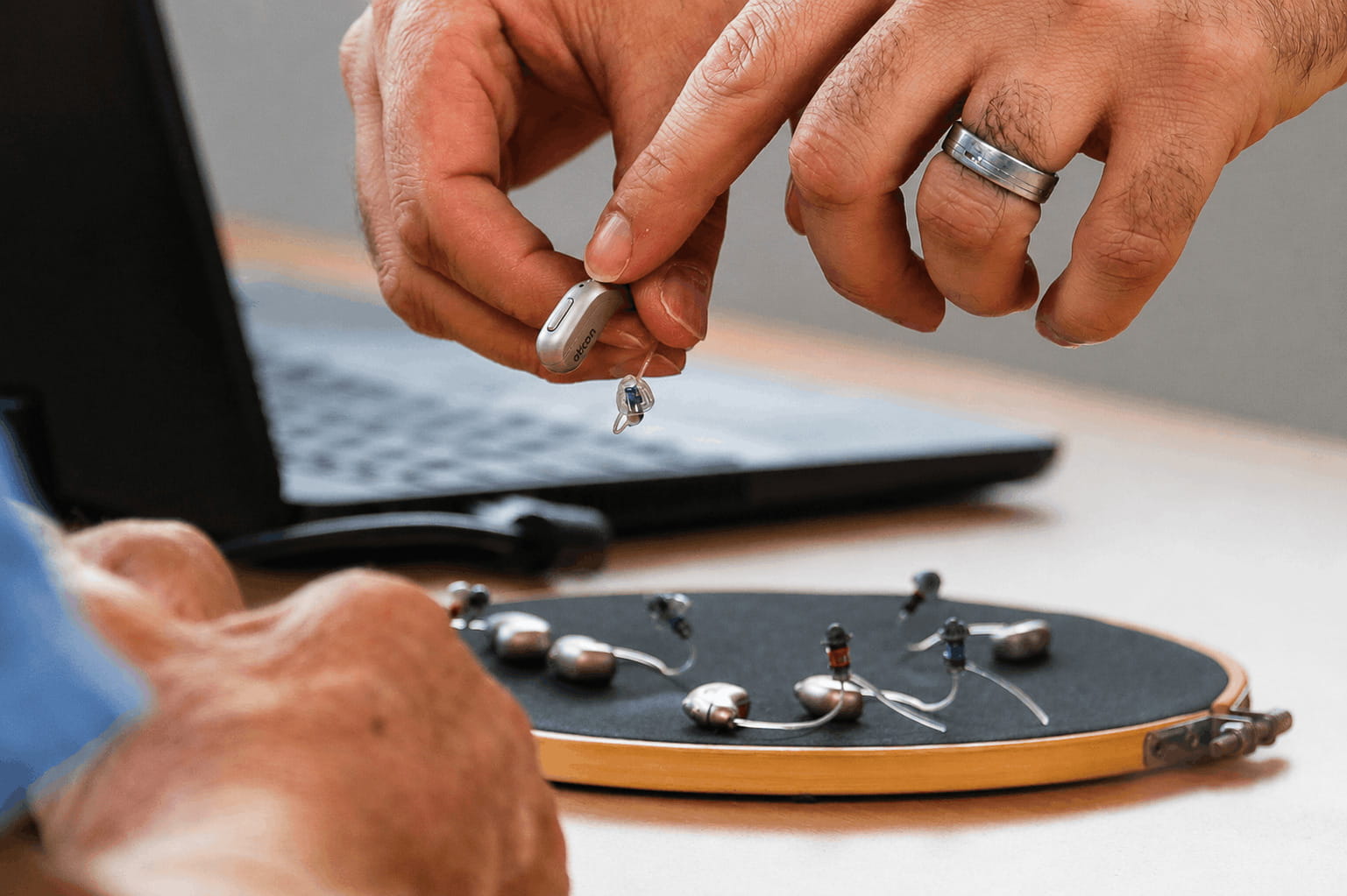 Close-up of hands manipulating a small, silver hearing aid with an attached clear tube, with a laptop in the background and several other hearing aid components arranged on a black surface within a wooden embroidery hoop.
