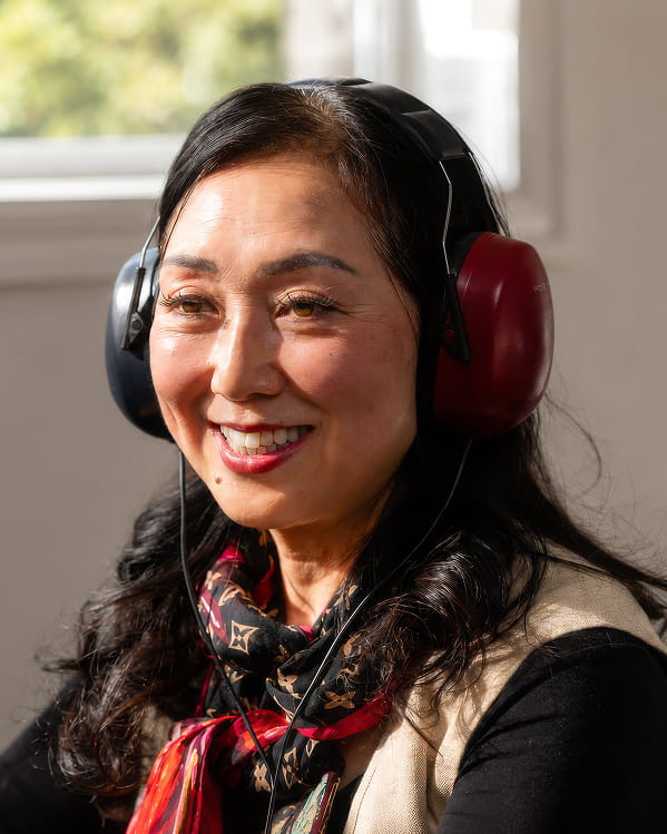 Close-up portrait of an Asian woman wearing large, dark headphones over her ears. She has long, dark wavy hair, and is smiling warmly. She wears a patterned scarf around her neck, and a light-colored vest over a dark garment. Sunlight illuminates her face from the side.