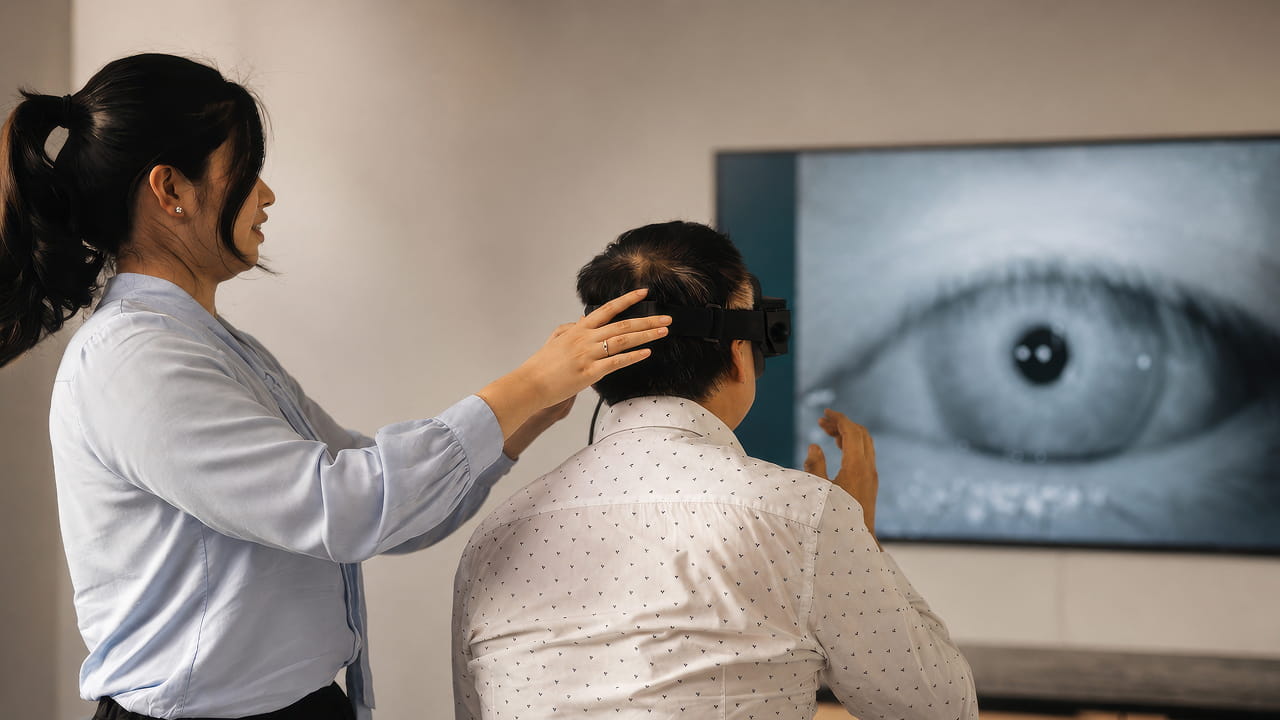 Man wearing a VR headset looks at a large screen displaying a close-up of an eye while a person adjusts the headset.