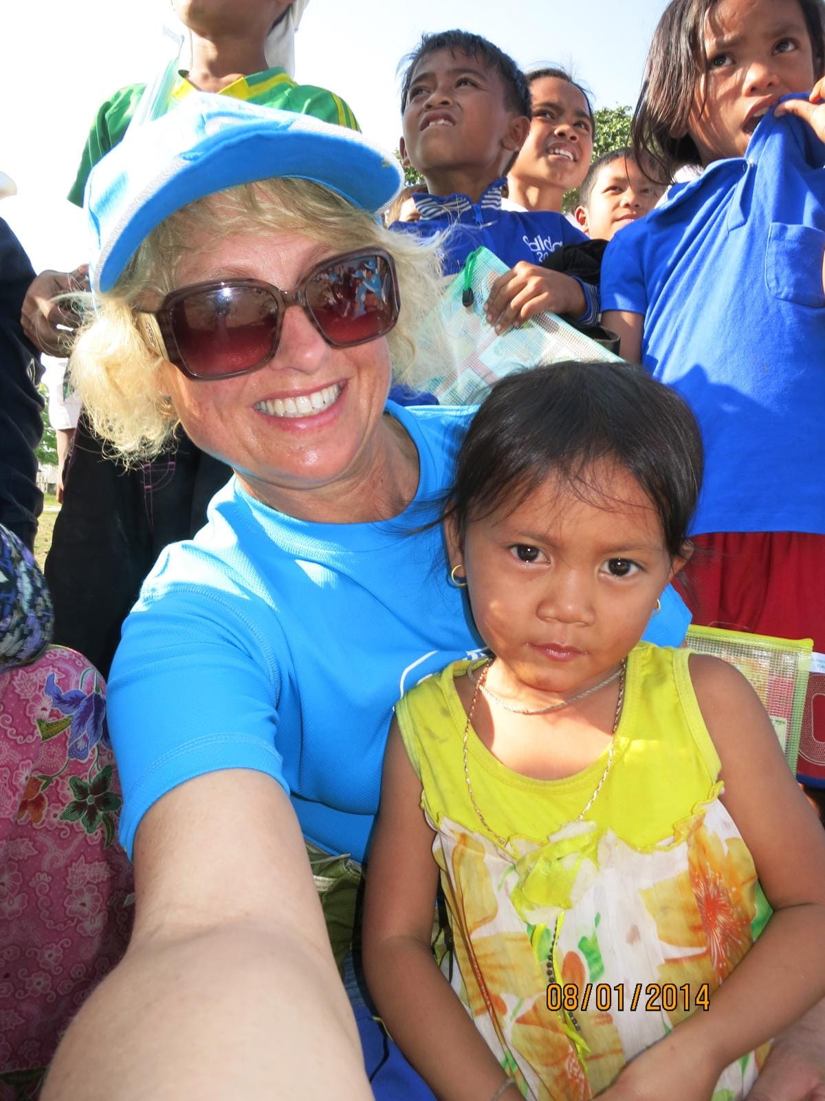 A smiling woman wearing sunglasses and a blue cap takes a selfie with a young girl in a yellow and white floral dress, surrounded by children in blue shirts.