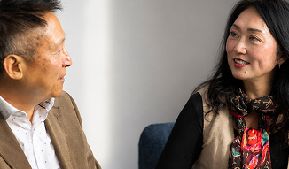 A man wearing a brown jacket and white patterned shirt faces a woman wearing a tan vest over a black shirt and a colorful patterned scarf, as they talk in a room.