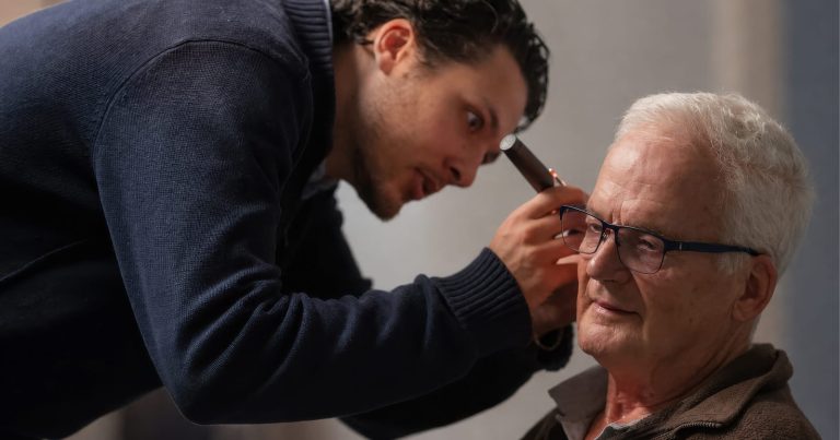 A man in a navy sweater leans forward to examine an older man's ear with an otoscope against a gray textured background.