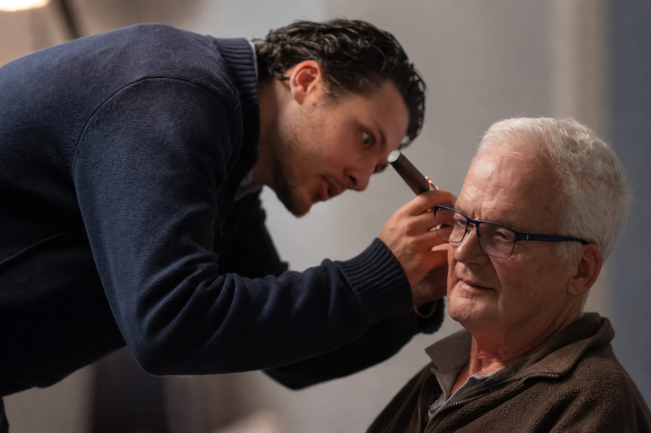 A man in a navy sweater leans forward to examine an older man's ear with an otoscope against a gray textured background.