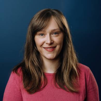 A woman wearing a pink sweater stands against a solid blue background, smiling confidently.