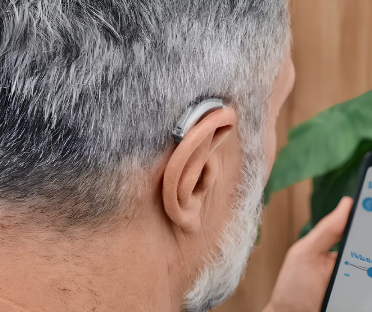 Close-up of a silver and grey hearing aid fitted behind the ear of a person with grey hair and a grey beard, who is holding a smartphone with an app open.