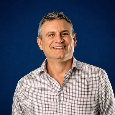 A man wearing a checkered shirt smiles warmly at the camera, showcasing a friendly demeanor.