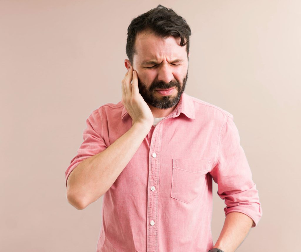 A man in a pink collared shirt winces with his eyes closed and hand pressed to his ear, indicating ear pain.