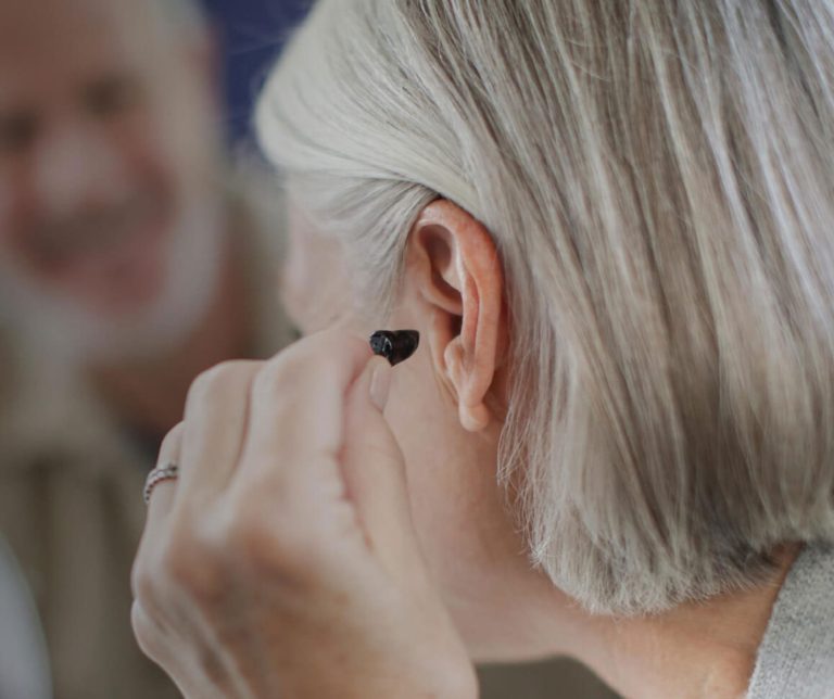 Close-up of a person inserting a small, dark hearing aid into their ear, with another person blurred in the background.