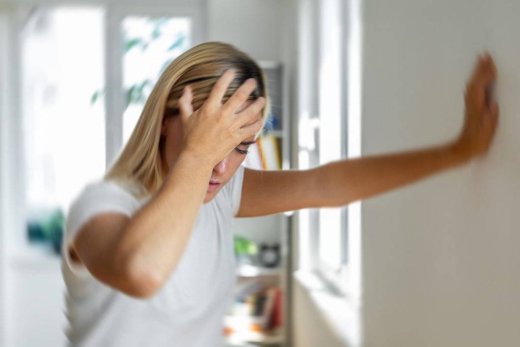 A woman stands in front of a wall, holding her head with a pensive expression, conveying a sense of contemplation or distress.